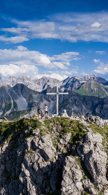 Mountain summit with cross and hikers, above the Alps, on a clear day with blue sky and clouds. | © Kleinwalsertal Tourismus eGen • Photographer: Bastian Morell