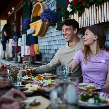 Ein glückliches Paar sitzt bei einem Sommeressen im Alpe Kuhgehren im Kleinwalsertal, umgeben von Dekorationen und Blumen vor einem holzverkleideten Gebäude. | © Kleinwalsertal Tourismus eGen • Fotograf: Oliver Farys
