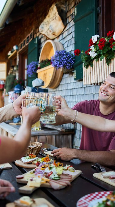 Menschen beim Sommer-Outdooressen auf einer Terrasse mit Holzhäuschen und bunten Blumen, Toasts und Snacks werden ausgetauscht. | © Kleinwalsertal Tourismus eGen • Fotograf: Oliver Farys