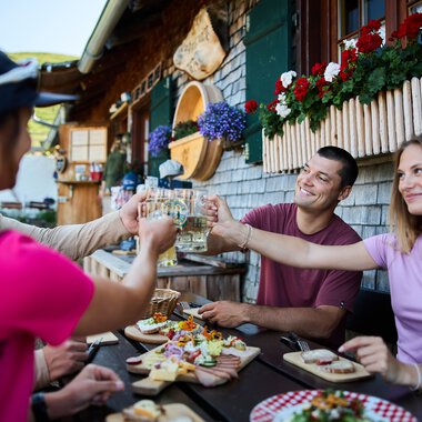 Menschen beim Sommer-Outdooressen auf einer Terrasse mit Holzhäuschen und bunten Blumen, Toasts und Snacks werden ausgetauscht. | © Kleinwalsertal Tourismus eGen • Fotograf: Oliver Farys