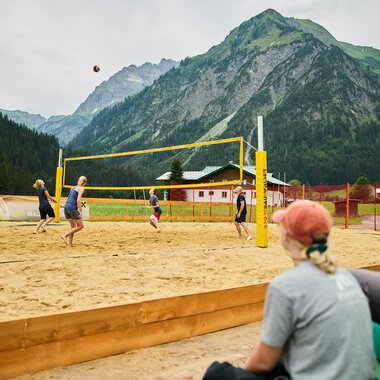 Menschen spielen Sommer-Volleyball auf einem Sandplatz vor einer bergigen Landschaft mit grünen Bergen und Wald im Hintergrund. | © Kleinwalsertal Tourismus eGen • Fotograf: Oliver Farys
