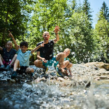 Kinder und ein Erwachsener spielen und springen in einem Fluss im Sommer, umgeben von grünen Bäumen im Kleinwalsertal. | © Kleinwalsertal Tourismus eGen • Fotograf: Oliver Farys