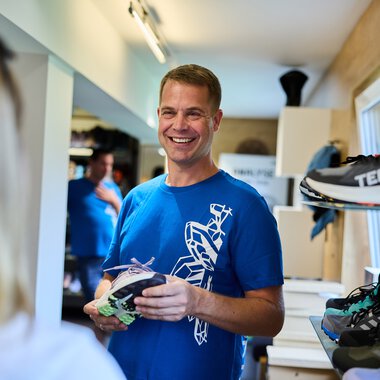 Ein lächelnder Mann im blauen T-Shirt in einem Geschäft, beim Verkauf von Sportschuhen. Im Hintergrund weitere Kunden und Regale mit Schuhen. | © Kleinwalsertal Tourismus eGen • Fotograf: Oliver Farys