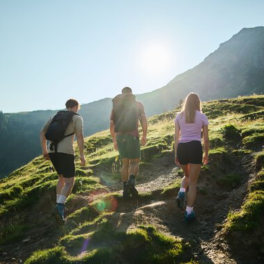 Drei junge Menschen wandern auf einem Waldweg im Kleinwalsertal bei Sonnenlicht, umgeben von grünen Hügeln und Bergen. | © Kleinwalsertal Tourismus eGen • Fotograf: Oliver Farys