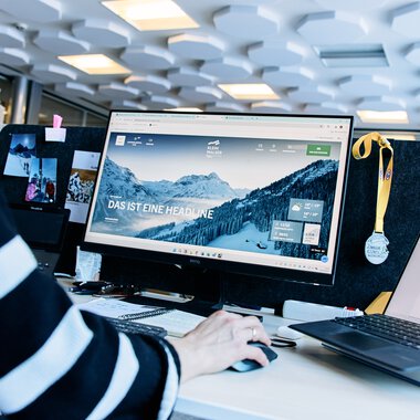 Arbeitsplatz in einem Büro mit Computer- und Laptop-Bildschirm, Medaille und Dokumenten, vermutlich während eines Meetings oder einer Arbeitssitzung. | © Kleinwalsertal Tourismus eGen • Fotograf: Oliver Farys