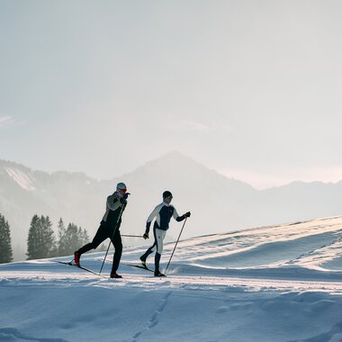 Zwei Langläufer beim klassischen Skilanglauf auf der Schwarzwasserloipe im Kleinwalsertal während des Winters, mit verschneiter Landschaft und Bergen im Hintergrund. | © Kleinwalsertal Tourismus eGen • Fotograf: Oliver Farys