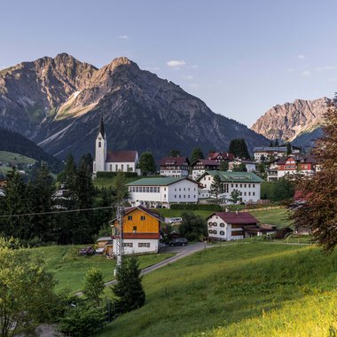Panoramaaufnahme eines kleinen Dorfes in den Bergen mit Kirche, umgeben von grünen Wiesen und Bäumen, vor einer dramatischen Bergkulisse. | © Kleinwalsertal Tourismus eGen • Fotograf: Steffen Berschin 