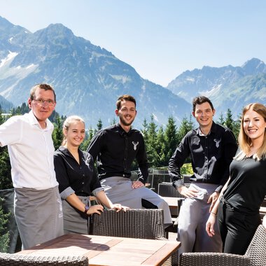 Fünf Mitarbeiter des Walserhofs sitzen auf einer Terrasse vor einer Bergkulisse mit Bergen und Wäldern, im sonnigen Wetter. | © Kleinwalsertal Tourismus eGen • Fotograf: Werner Krug