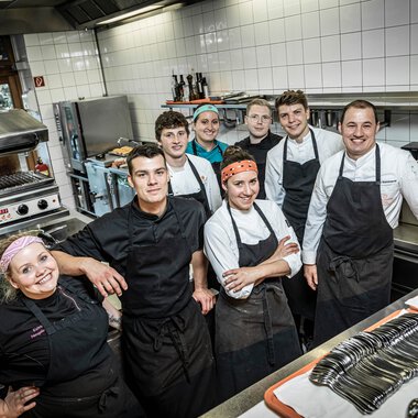 Team aus Küchenmitarbeitern in einer Restaurantküche, alle in Arbeitskleidung und aprons, bereit für den Service. | © Kleinwalsertal Tourismus eGen • Fotograf: Werner Krug