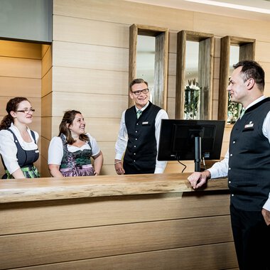 Mitarbeiter an der Rezeption eines Hotels, freundlich im Gespräch, mit Computer und Blumen im Hintergrund. | © Kleinwalsertal Tourismus eGen • Fotograf: Werner Krug