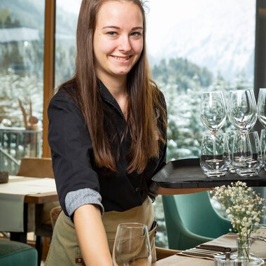 Eine junge Frau mit langen braunen Haaren, lächelnd, steht an einem Tisch in einem Restaurant mit großen Fenstern und Blick auf die Berge. | © Kleinwalsertal Tourismus eGen • Fotograf: Werner Krug