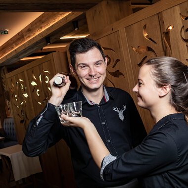 Ein junger Mann und eine junge Frau in einem Restaurant, die sich freundlich zuprosten, der Mann hält eine Weinkugel, die Frau reicht ihm ein Weinglas. | © Kleinwalsertal Tourismus eGen • Fotograf: Werner Krug