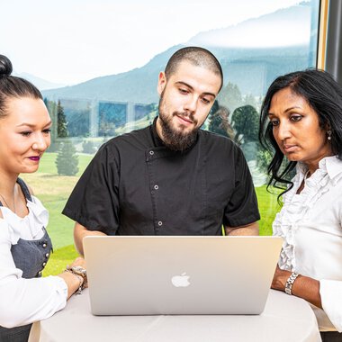 Drei Mitarbeitende schauen auf einen Laptop während einer Besprechung vor einer großen Fensterfront mit Bergblick. | © Kleinwalsertal Tourismus eGen • Fotograf: Werner Krug
